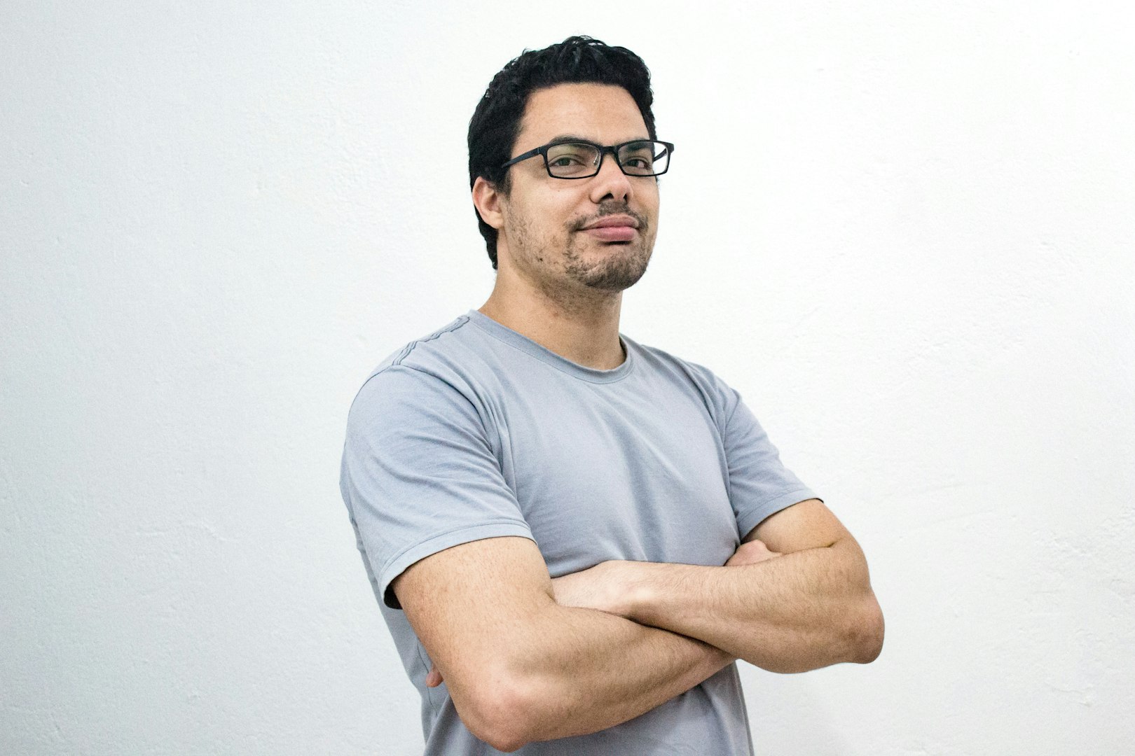 A man with wavy brown hair and a beard is standing in front of a shelf filled with various household items. He is wearing a light blue polo shirt and has a neutral expression.